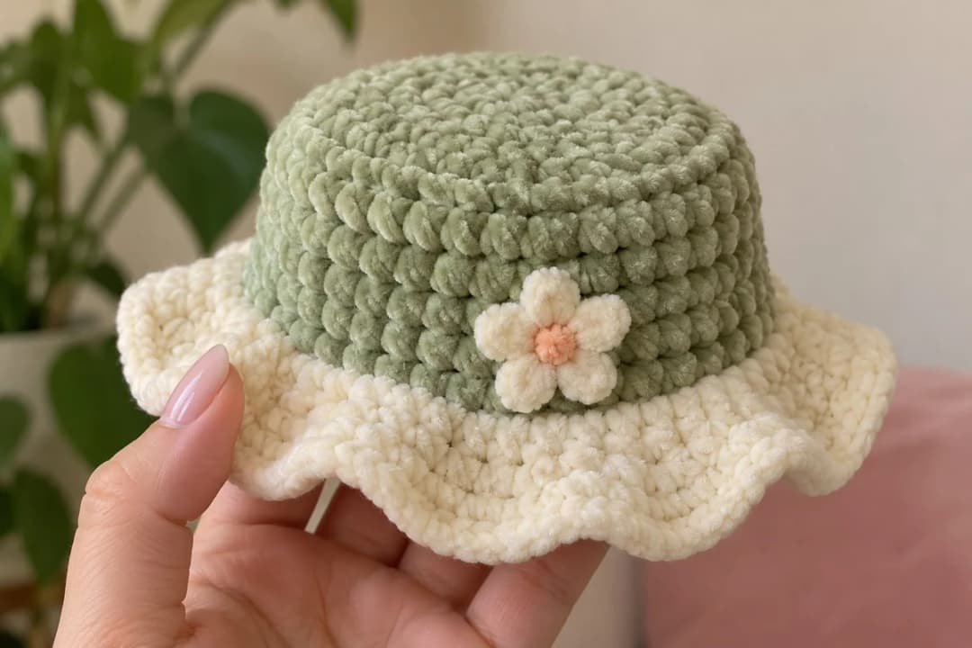 The sage green gardener's hat held in one hand: a round chenille crown with straight sides, a small cream daisy with an apricot center embroidered just above the brim, and a wide cream brim falling in soft waves all around