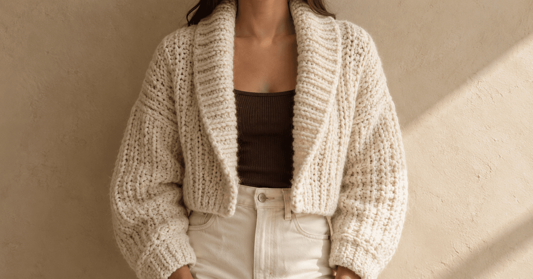 A woman wearing a handmade chunky crochet cardigan in cream super-bulky wool — cropped waist-length, oversized drop-shoulder fit, with a wide shawl collar folded outward and tall knit-look vertical column stitches on the body and sleeves. Underneath, a chocolate brown ribbed tank and high-waist cream jeans. Soft beige plaster wall background with a warm diagonal window-light shadow from the side.