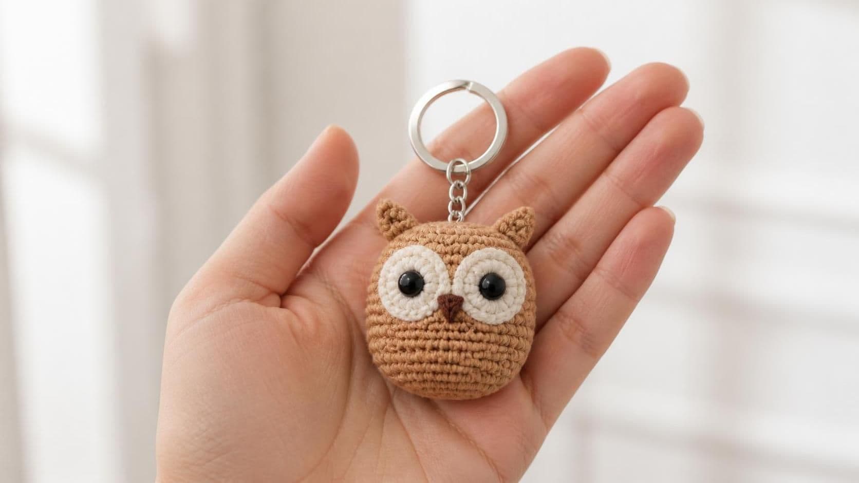 A handmade mini crochet owl keychain held in the palm of a hand — a small round tan-brown owl amigurumi about 5 cm tall with large black safety eyes, cream face disc, tiny brown beak and two little ear tufts, on a silver split-ring keychain in soft indoor light against a white door