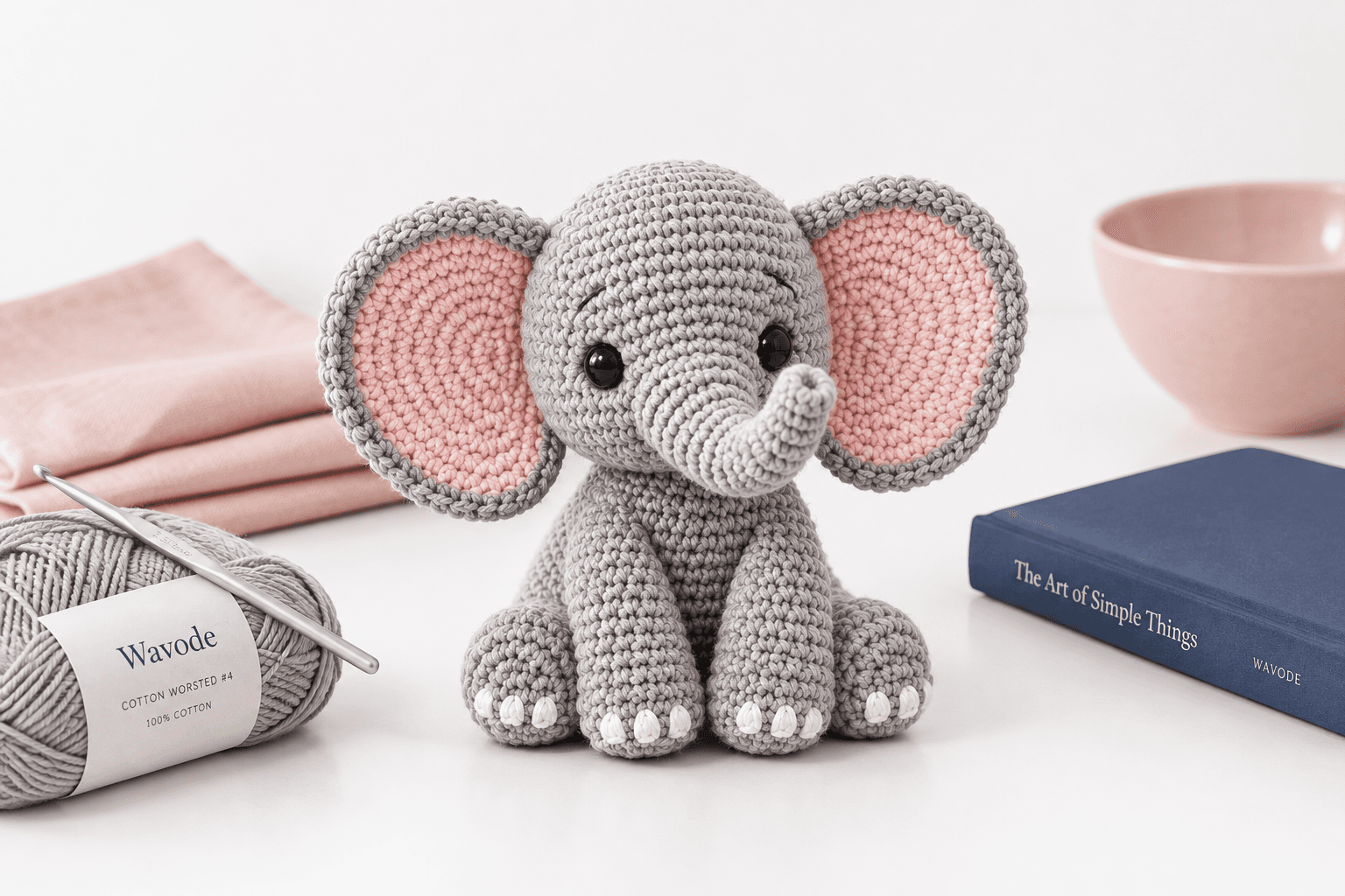 Grey crochet elephant amigurumi with big pink-lined ears, a curled-up trunk, black safety eyes and tiny white toenails, sitting on a pure white surface with a blush linen napkin, a navy hardcover book, and a Wavode grey worsted cotton skein beside it