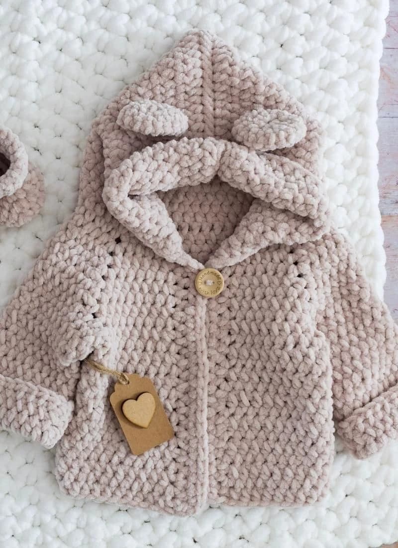 A dusty pink plush chenille crochet kids hoodie with two round bear ears, a folded-back hood brim, one wooden "handmade with love" button at the neck and turned-up cuffs, laid flat on a white chunky blanket with a kraft heart tag