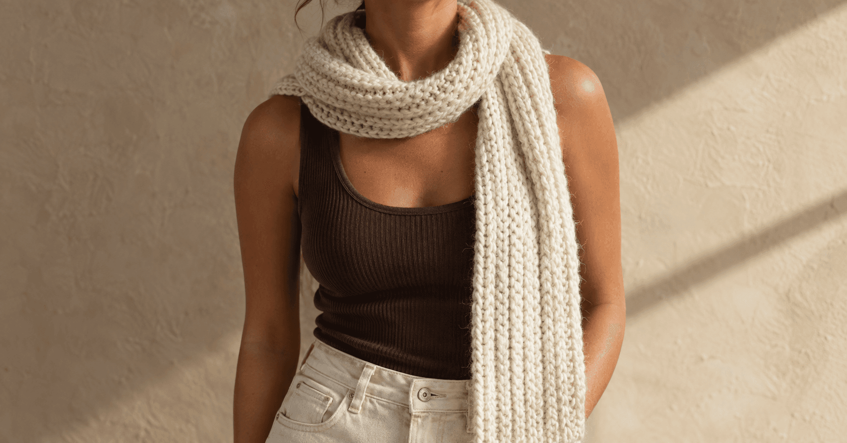 A woman wearing a handmade cream Tunisian-crochet ribbed wrap scarf loosely draped around her neck, with one long ribbed end trailing down her chest and the other tucked behind her shoulder. Distinct vertical 2x2 rib columns in bulky cream merino wool. Underneath, a chocolate brown ribbed cotton tank and cream jeans. Soft beige plaster wall background with warm diagonal window-light shadow from the side.
